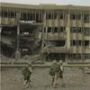 Marines in combat gear and holding weapons walk through a patch of sand and rocks in front of a heavily damaged building.