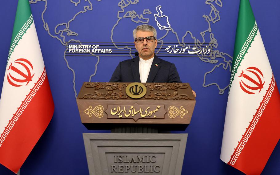 Baghaei speaks at a lectern between two Iran flags.