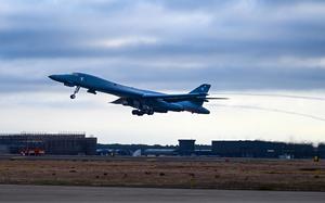 A U.S. Air Force strategic bomber takes off from an airfield under a cloudy sky. The aircraft is slightly tilted upward as it ascends, with landing gear still extended and heat distortion visible from its engines. In the background, airport infrastructure, including hangars and a control tower, is seen on the tarmac surrounded by grassy terrain.