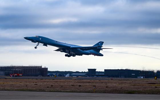 A U.S. Air Force strategic bomber takes off from an airfield under a cloudy sky. The aircraft is slightly tilted upward as it ascends, with landing gear still extended and heat distortion visible from its engines. In the background, airport infrastructure, including hangars and a control tower, is seen on the tarmac surrounded by grassy terrain.