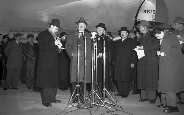 A black and white photo of a man speaking in front of a microphone, surrounded by others, some of whom appear to be reporters.