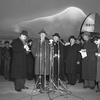 A black and white photo of a man speaking in front of a microphone, surrounded by others, some of whom appear to be reporters.