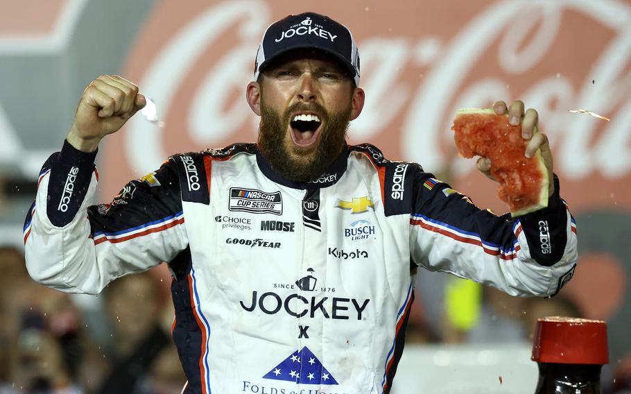 A NASCAR driver in a white racing suit with red, white and blue trim celebrates enthusiastically with both fists raised, shouting with joy. 