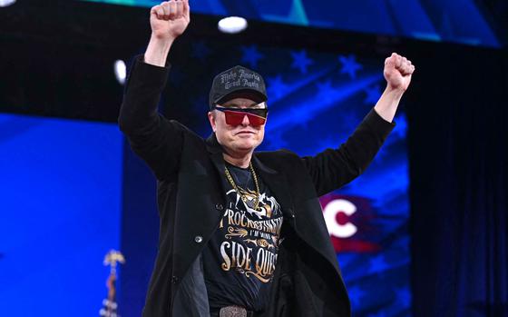 Elon Musk wears a “Make America Great Again” cap, red-tinted sunglasses, a gold chain and a black coat while raising both fists in the air and standing on a stage. He is also wearing a black T-shirt with a graphic and text that reads “I’m not procrastinating, I’m doing side quests.”