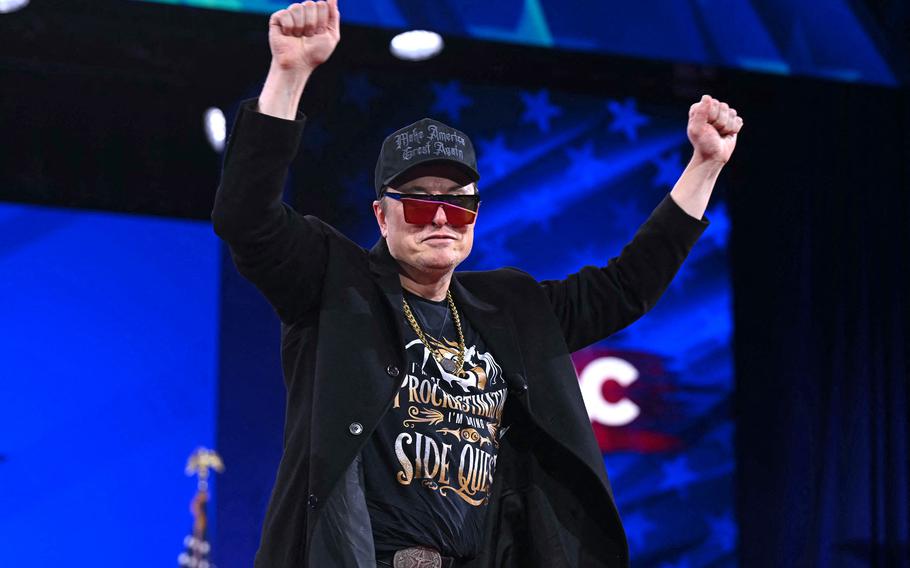 Elon Musk wears a “Make America Great Again” cap, red-tinted sunglasses, a gold chain and a black coat while raising both fists in the air and standing on a stage. He is also wearing a black T-shirt with a graphic and text that reads “I’m not procrastinating, I’m doing side quests.”