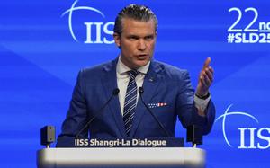 Pete Hegseth gestures with his left arm outstretched as he stands at a podium and speaks into microphones on a stage in front of a large blue screen.