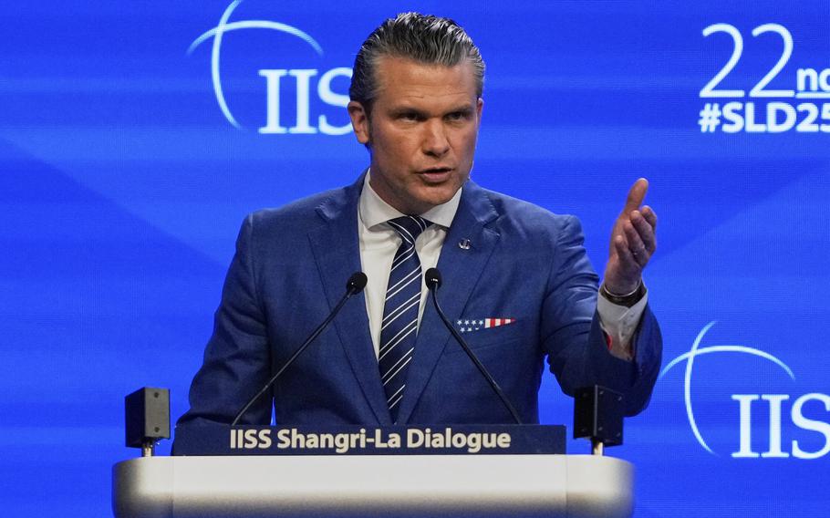 Pete Hegseth gestures with his left arm outstretched as he stands at a podium and speaks into microphones on a stage in front of a large blue screen.