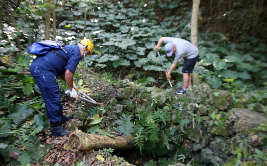 A member of the Japan Maritime Self-Defense Force and a U.S. Navy sailor work to clear jungle brush growing up through the Chibu nu Ka well, a sacred fertility site at White Beach Naval Facility, Okinawa, Friday, Feb. 17, 2023.