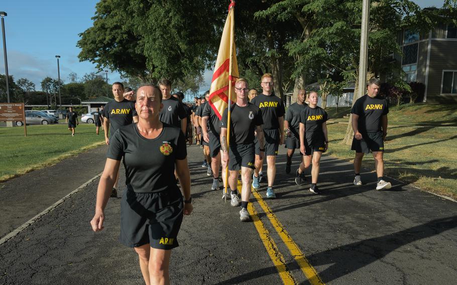 A group of soldiers in black shirts and shorts walk along a road.