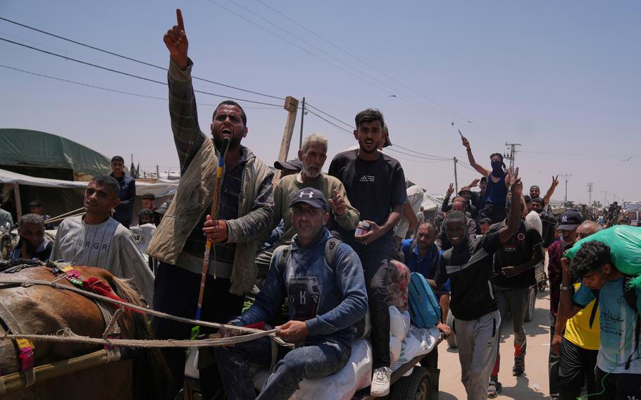 Palestinians ride in a line of carts and walk next to them on a dirt road carrying bags of food and humanitarian aid 