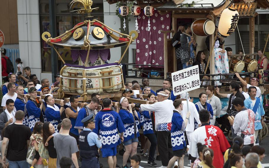 A large crowd of festival participants, many wearing blue and white traditional Japanese festival clothing, carry a decorated portable shrine (mikoshi) through a busy street. The ornate shrine features golden decorative elements, purple fabric and traditional symbols. Spectators line the sides of the street, and there are traditional festival banners and decorations visible in the background.