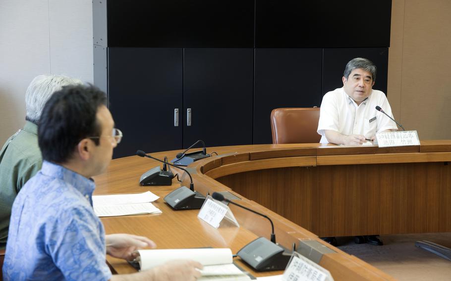 Three men sit at a conference table with microphones. Two men, one in a blue shirt and one in a green shirt, are turned toward a man in a white shirt who is speaking into a microphone. 