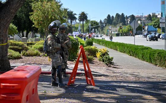 Two soldiers in camouflage uniforms and helmets and carrying weapons stand on a street next to an orange barrier, with a tree-lined sidewalk behind them.