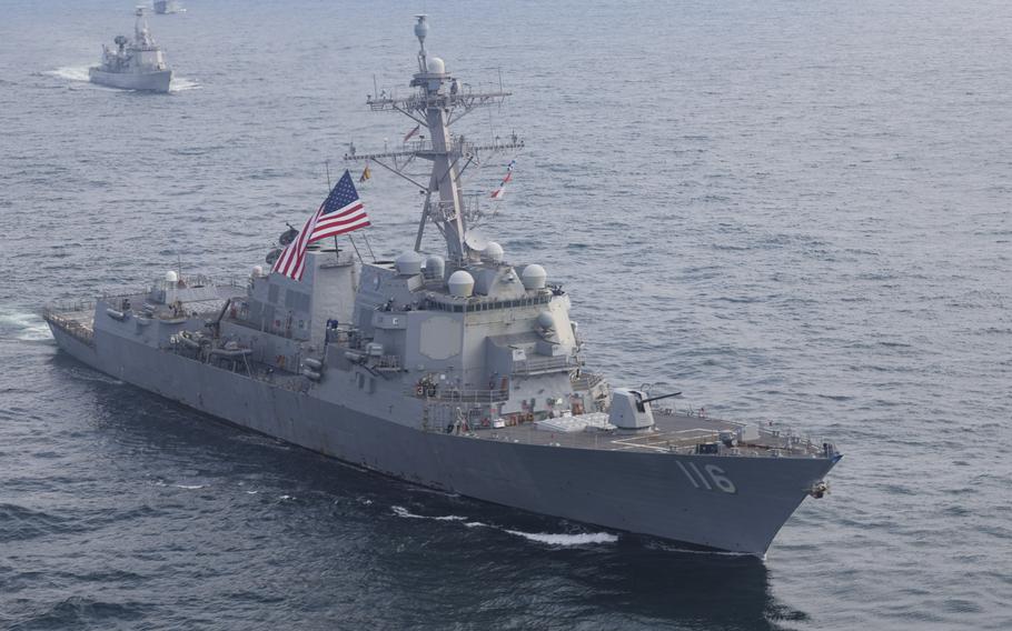 A naval destroyer flying a U.S. flag sails in open ocean with other ships and the horizon in the background.