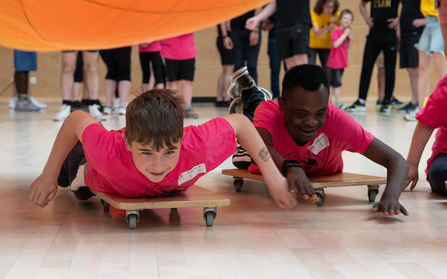 Two children ride scooter boards on their stomachs underneath an orange playground parachute. 