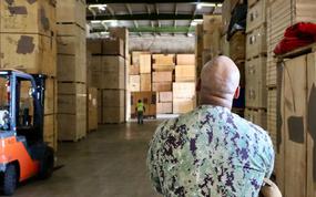 A man in camouflage military uniform, seen from behind, stands in a warehouse between large stacks of pallets and cardboard boxes.