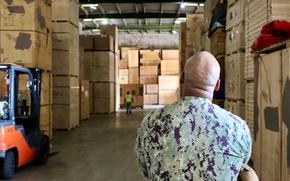 A man in camouflage military uniform, seen from behind, stands in a warehouse between large stacks of pallets and cardboard boxes.