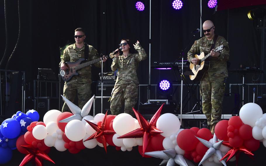 A woman in camouflage uniform and sunglasses sings into a microphone on a stage, while two men in camouflage uniforms and sunglasses play guitars on either side of the woman. A large strand of red, white and blue balloons runs the edge of the stage at their feet.