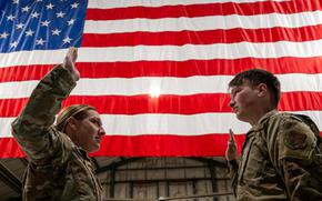 An airman raises her hand to reenlist another service member in front of a giant American flag.