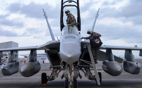 Two military airmen in flight suits and helmets climb out of a fighter jet, seen from the front.