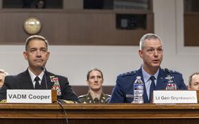 A Navy officer and an Air Force officer, both in dress uniforms, sit at a table in front of microphone in a congressional hearing room.