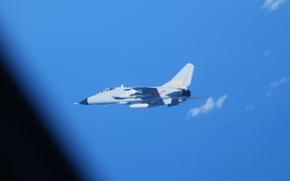A military fighter aircraft flies against a bright blue sky with scattered white clouds.