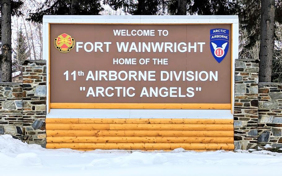 A brown and yellow entrance sign reading “Welcome to Fort Wainwright” with snow on the ground beneath it and trees behind.