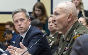 An Army officer in dress uniform speaks into a microphone while seated a table in a congressional hearing room, while a man in a dark suit looks on from the left.