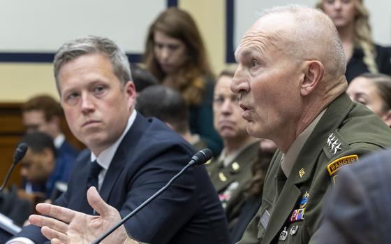 An Army officer in dress uniform speaks into a microphone while seated a table in a congressional hearing room, while a man in a dark suit looks on from the left.