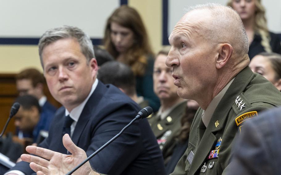 An Army officer in dress uniform speaks into a microphone while seated a table in a congressional hearing room, while a man in a dark suit looks on from the left.