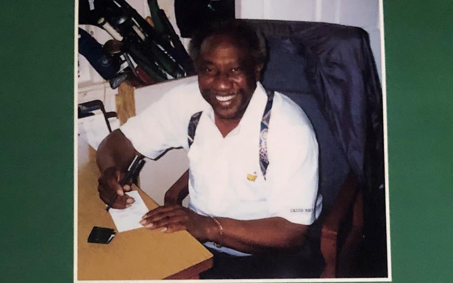 A man seated at a desk looks up and smiles while holding a pen over a piece of paper.