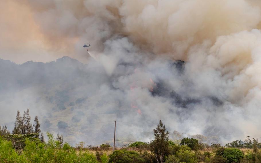 A helicopter, seen from a distance, drops water from a red bucket hanging underneath it on a fire burning a forested area as large plumes of smoke billow into the sky and obscure the view.