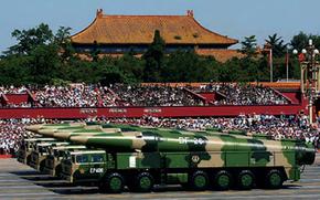 Camouflage military vehicles carrying large camouflage missiles drive past a grandstand full of people, with the roof of a Chinese building seen above trees in the background.