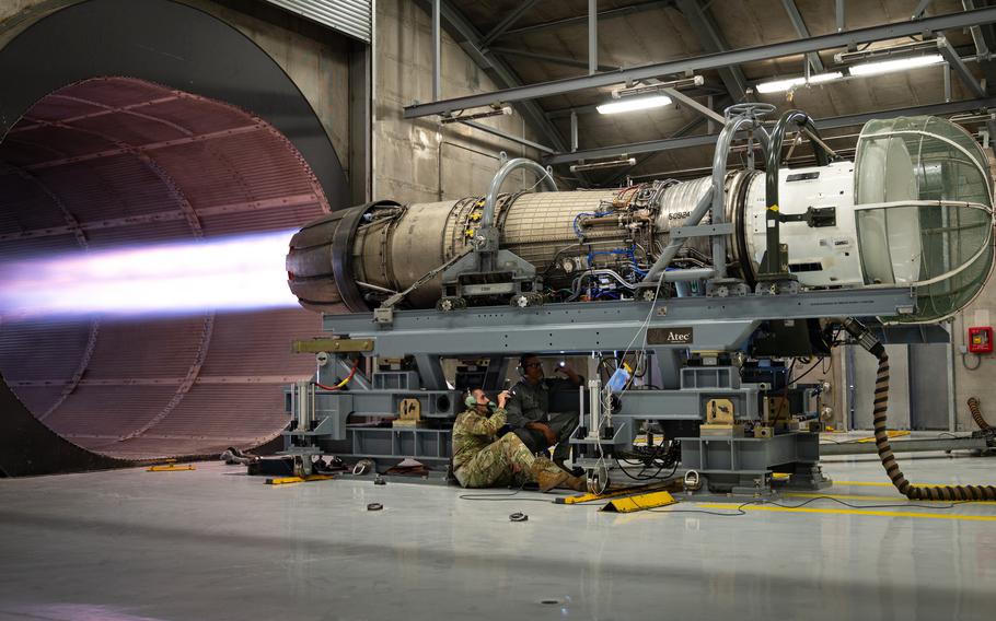 A service member sit near an engine that is blasting fire into a tunnel.