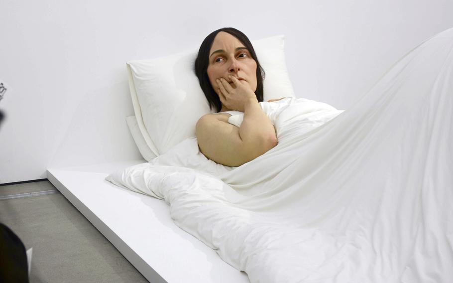 A large, life-like sculpture of a woman in bed with her head against a pillow, a white sheet over her body and her hand on her chin rests of a white platform in a museum.