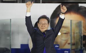 A South Korean man in a dark suit and glasses raises his arms in celebration on a stage in front of a large photo of himself.
