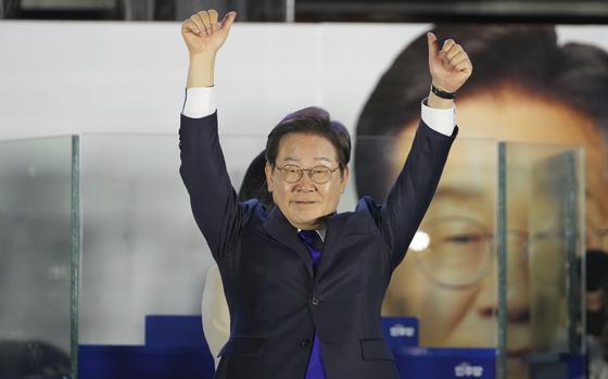 A South Korean man in a dark suit and glasses raises his arms in celebration on a stage in front of a large photo of himself.