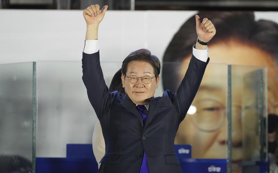 A South Korean man in a dark suit and glasses raises his arms in celebration on a stage in front of a large photo of himself.