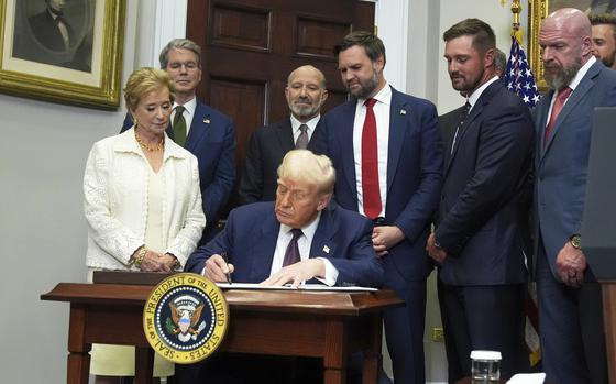 Donald Trump sits at a wooden table adorned with the presidential seal and signs a document, while administration officials stand behind him.
