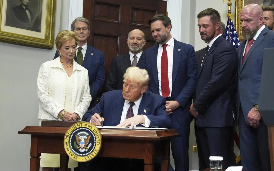Donald Trump sits at a wooden table adorned with the presidential seal and signs a document, while administration officials stand behind him.