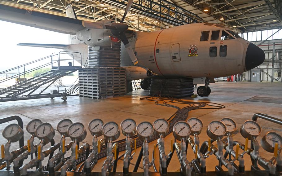  In the foreground, numerous pressure gauges are arranged in rows on an orange testing platform, with cables and hoses connecting to a military aircraft’s undercarriage. The aircraft sits on multiple rows of black inflatable air bags.
