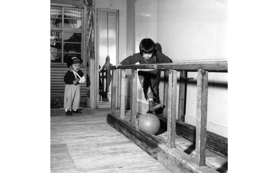 A young Korean child strengthens their muscles weakened by polio by kicking a medicine ball the length of a walking rack at the National Rehabilitation Center in Pusan [Busan]. Looking on is an even younger patient at the hospital with an arm injury. The mirror in the background allows the polio sufferers to observe their own progress. 