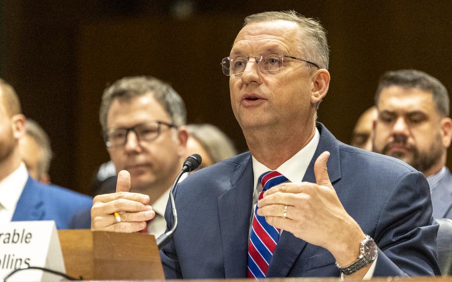 Doug Collins in a suit and red and blue tie, gesturing with both hands while seated and speaking before a microphone.