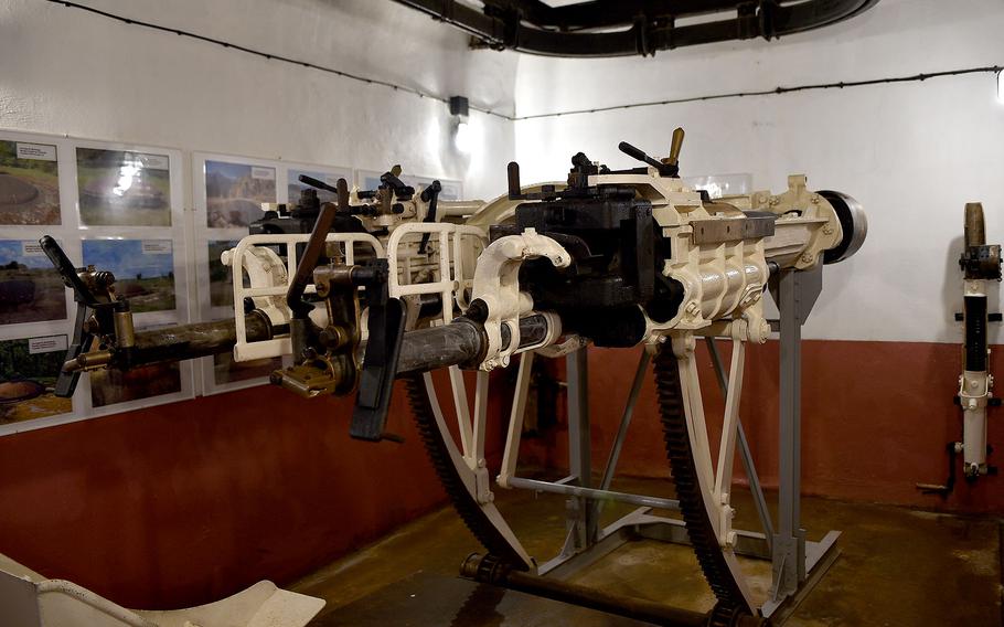 Interior museum display showing a mounted machine gun on a white stand, with informational posters and photographs on the walls.