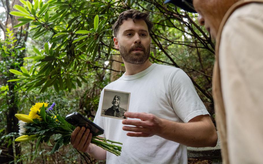 A man in a white T-shirt holds up a black-and-white photo of an aviator in one hand while holding flowers and a phone in the other, with a forest scene behind him.