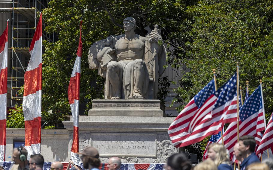 Marchers go past the statue of a seated man. The inscription says “Eternal viligance is the price of liberty.”