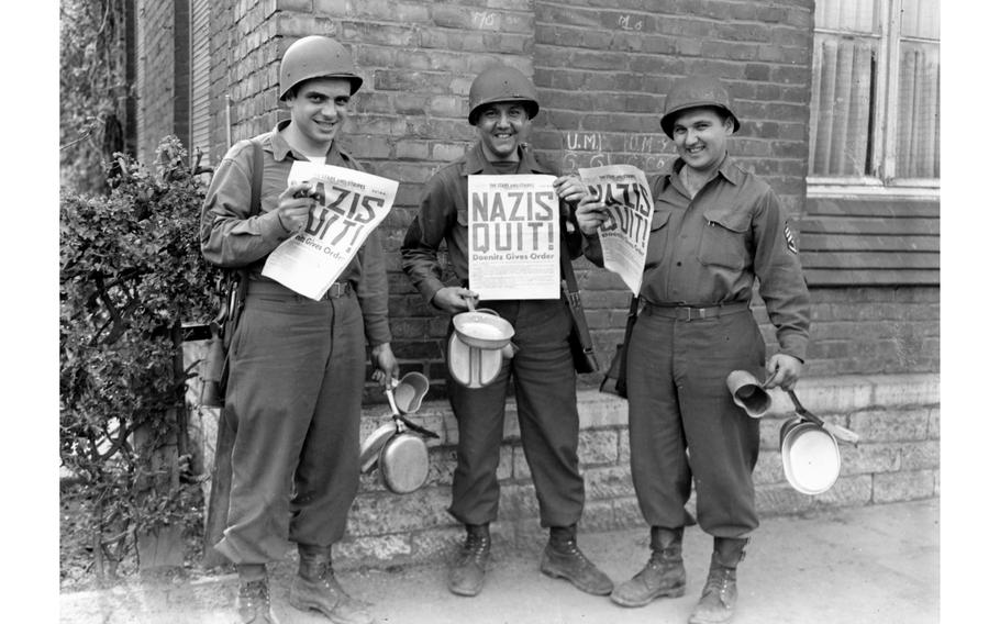 A black and white photo of three soldiers in 1940s Army uniforms holding newspapers with the headline “NAZIS QUIT!”