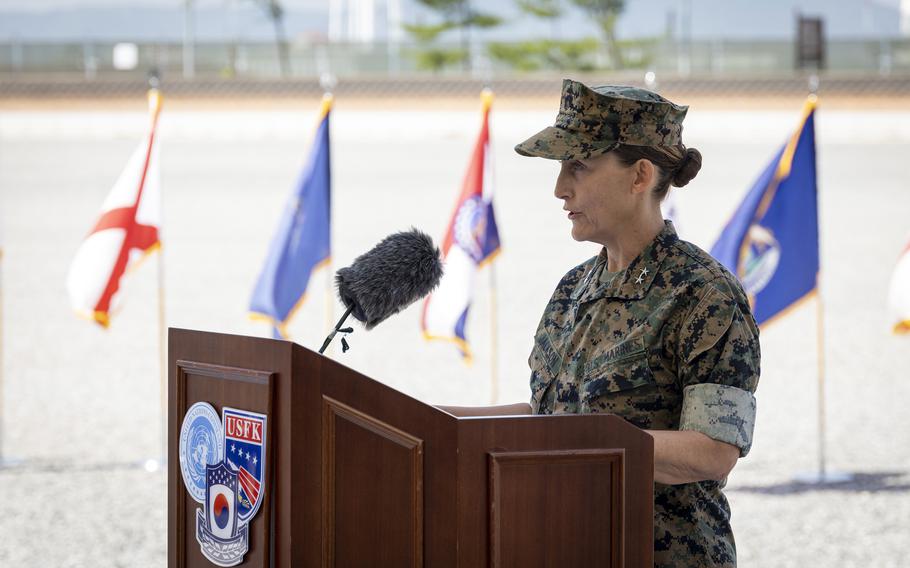 A female Marine officer in digital woodland camouflage uniform speaks at a wooden podium in an outdoor setting. The podium displays the United States Forces Korea emblem. 