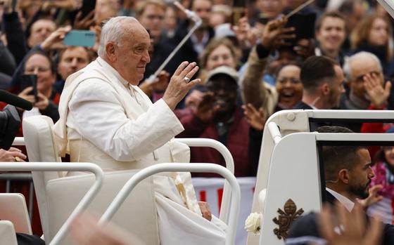 Pope Francis, dressed in white, waves to a cheering crowd from the Popemobile as people take photos and reach out toward him.
