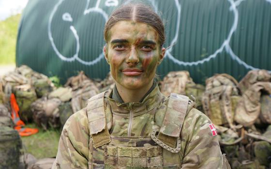 A woman wearing a camouflage military uniform and camouflage face paint looks forward while posing in front of a row of camouflage duffel bags on the ground.